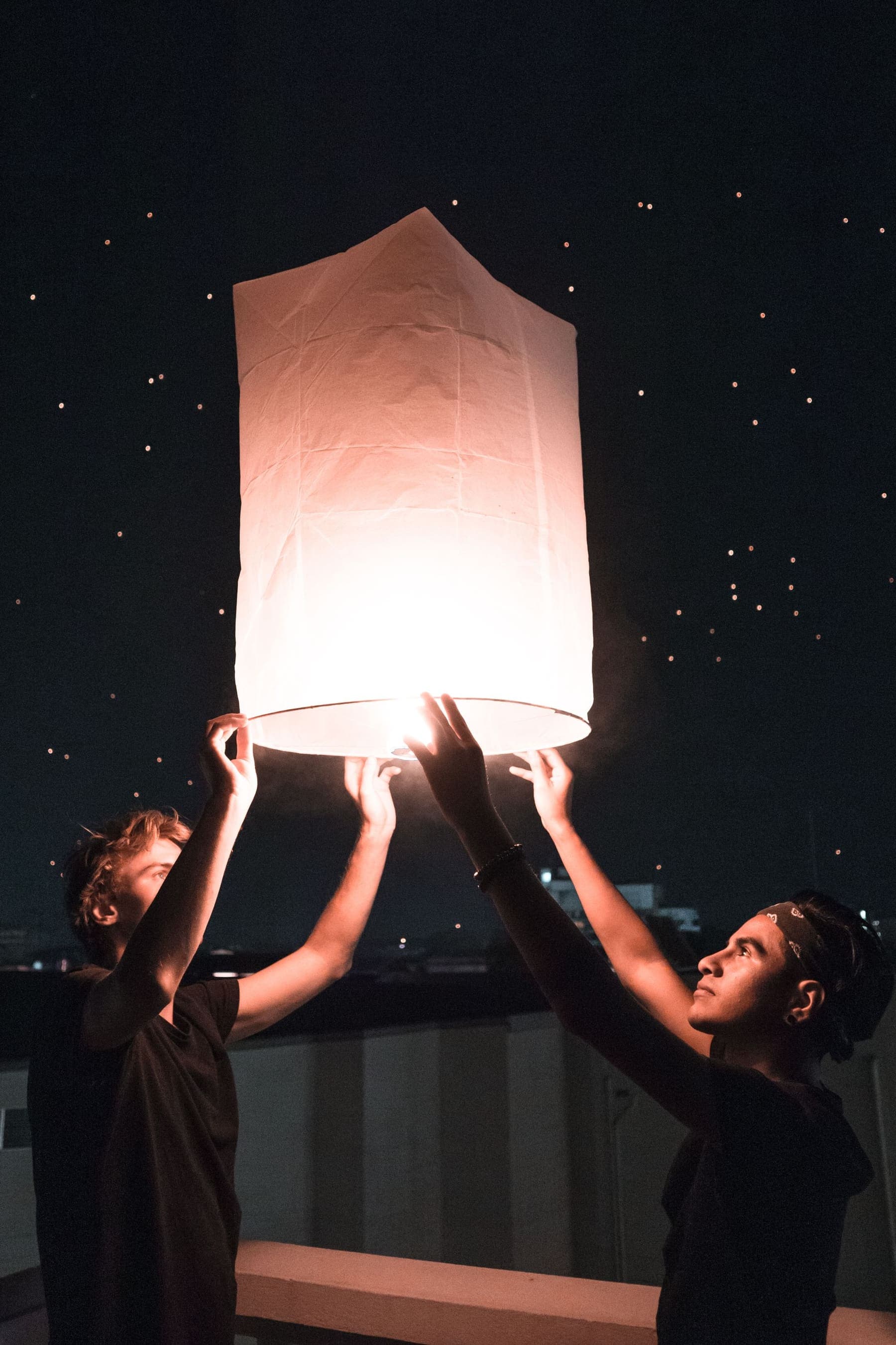 Two people release a glowing paper sky lantern at night in Chiang Mai, with more lanterns in the sky